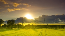 A row of wind turbines stand in a country field against a partly cloudy sky with the sun setting behind the clouds.