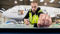 A young male technician wearing a high-visibility jacket stands in an aircraft hangar with a Lufthansa aircraft in the background and carefully applies sharkskin film to a flat surface.
