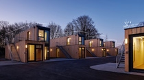A small village of wooden-cladded tiny houses with large windows made from upcycled shipping containers stacked one on top of another. It is dusk and warm-colored lights are illuminating the interiors. (Photo: Containerwerk) A small village of wooden-cladded tiny houses with large windows made from upcycled shipping containers stacked one on top of another. It is dusk and warm-colored lights are illuminating the interiors. (Photo: Containerwerk)