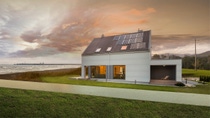 Stormy dark clouds gather over a white modern house with a small green lawn and solar roof panels located next to a beach with the rough waves just a few meters away.