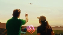 A male and female farmer are standing outdoors in South America, facing away from the camera, observing a drone flying in the sky. The man is holding a tablet and points towards the drone. The woman is looking at the drone. A light swirl hovers between them. A male and female farmer are standing outdoors in South America, facing away from the camera, observing a drone flying in the sky. The man is holding a tablet and points towards the drone. The woman is looking at the drone. A light swirl hovers between them.