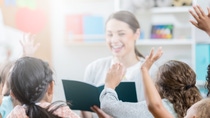 A group of unrecognizable schoolchildren sit on the floor of their classroom and raise their hands for their smiling teacher.  The teacher is elevated as she holds an open book and talks to them.