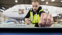 A young male technician wearing a high-visibility jacket stands in an aircraft hangar with a Lufthansa aircraft in the background and carefully applies sharkskin film to a flat surface.