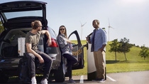 A group of three people, two men and one women, is chatting casually and drinking coffee, while their EV is charging at a charging station. In the backdrop, one can see green hills and some windmills. 