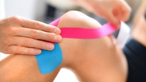 Kinesiology taping treatment with blue and pink tape on athlete patient injured knee. Woman hands apply kinesio treatment after sports muscle injury.