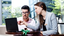 A man and a woman in a suit sitting next to each other looking at a laptop and talking