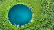 Aerial view of a circular deep‑blue lake surrounded by dense green vegetation. Aerial view of a circular deep‑blue lake surrounded by dense green vegetation.