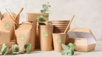 The image shows a variety of eco-friendly food packaging products made from brown, biodegradable materials. The packaging is branded with the "ecovio" logo. Green leaves are scattered among the items. The image shows a variety of eco-friendly food packaging products made from brown, biodegradable materials. The packaging is branded with the "ecovio" logo. Green leaves are scattered among the items.
