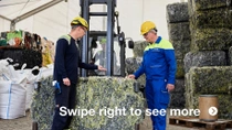 Two men in yellow helmets and work overalls stand beside a large, pressed block of textile waste. Two men in yellow helmets and work overalls stand beside a large, pressed block of textile waste.