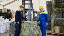 Two men in yellow helmets and work overalls stand beside a large, pressed block of textile waste. Two men in yellow helmets and work overalls stand beside a large, pressed block of textile waste.
