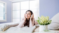 Foto: eine Frau mit Kopfhörern liegt auf einem Bett, im Vordergrund steht ein Strauß Maiglöckchen. Foto: eine Frau mit Kopfhörern liegt auf einem Bett, im Vordergrund steht ein Strauß Maiglöckchen.