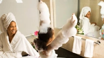 A cozy bathroom with a bathtub and a mirror. A smiling woman sits on the edge of a bathtub in a white bathrobe with a towel wrapped around their head. A girl is in the tub, covered in bubbles with arms raised. 