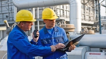 Two men wearing blue uniform and yellow hard hat looking at a laptop