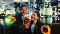 A man and a woman in casual jackets stand in front of an Asian city skyline at night. They are both looking at his phone. Light swirls appear next to them. A man and a woman in casual jackets stand in front of an Asian city skyline at night. They are both looking at his phone. Light swirls appear next to them.