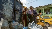 A man writing something in a notebook. A woman is standing next to him looking at the book. They are standing by a dumpster and there are bags of trash in front of them. A man writing something in a notebook. A woman is standing next to him looking at the book. They are standing by a dumpster and there are bags of trash in front of them.