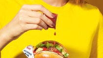 Frau hält einen Burger in der einen Hand während sie mit der anderen Hand den Burger mit Ketschup beträufelt. Frau hält einen Burger in der einen Hand während sie mit der anderen Hand den Burger mit Ketschup beträufelt.
