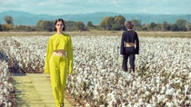 A fashion show is taking place in a cotton field, featuring models among the cotton plants. A woman in a bright yellow suit walks towards the camera. A fashion show is taking place in a cotton field, featuring models among the cotton plants. A woman in a bright yellow suit walks towards the camera.