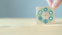 Four wooden blocks form a circular graphic with icons for recycling, sustainability, and production; a hand is aligning the blocks. Four wooden blocks form a circular graphic with icons for recycling, sustainability, and production; a hand is aligning the blocks.