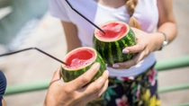Two people hold hollowed-out watermelons filled with a red beverage, each with a black straw. One person is not visible, the other is wearing a white shirt and floral-patterned pants. Two people hold hollowed-out watermelons filled with a red beverage, each with a black straw. One person is not visible, the other is wearing a white shirt and floral-patterned pants.