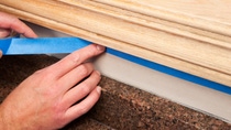 A painter is applying blue masking tape to protect an area of wall below oak window trim which is being refinished. The wall repainted after staining the trim, below the hand is a granite countertop. A painter is applying blue masking tape to protect an area of wall below oak window trim which is being refinished. The wall repainted after staining the trim, below the hand is a granite countertop.