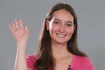 A young woman stands in front of a neutral background and smiles at the camera. She has raised her hand in a friendly greeting. A young woman stands in front of a neutral background and smiles at the camera. She has raised her hand in a friendly greeting.