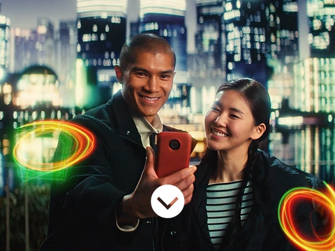 A man and a woman in casual jackets stand in front of an Asian city skyline at night. They are both looking at his phone. Light swirls appear next to them.