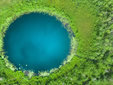Aerial view of a circular deep‑blue lake surrounded by dense green vegetation. Aerial view of a circular deep‑blue lake surrounded by dense green vegetation.