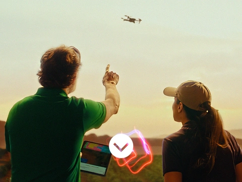  A male and female farmer are standing outdoors in South America, facing away from the camera, observing a drone flying in the sky. The man is holding a tablet and points towards the drone. The woman is looking at the drone. A light swirl hovers between them.