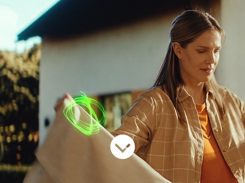 A woman is standing outdoors, taking a tablecloth down from a clothesline in front of a house and some greenery. There are light swirls near her hands holding the cloth.