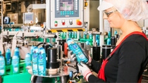 A female production worker wearing protective gloves, safety glasses and hairnet inspects a bottle of green-blue colored shower gel on a production line. 