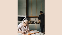 Older woman sitting at a table doing sudokus while a man is washing dishes in the background. Older woman sitting at a table doing sudokus while a man is washing dishes in the background.