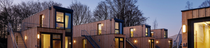 A small village of wooden-cladded tiny houses with large windows made from upcycled shipping containers stacked one on top of another. It is dusk and warm-colored lights are illuminating the interiors. (Photo: Stefan Hohloch for Containerwerk)