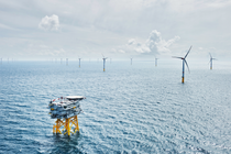 A wide view of a blue ocean with waves taken from above, showing two rows of wind turbines stretching into the distance and an offshore platform with helicopter landing pad in the foreground.