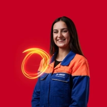 A female BASF employee in an orange and blue work overall standing against a red background, a light swirl hovers around her shoulder. A female BASF employee in an orange and blue work overall standing against a red background, a light swirl hovers around her shoulder.